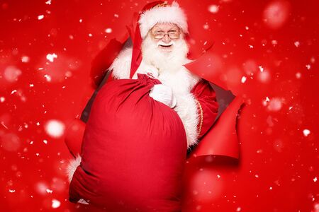 Santa Claus , the real one, comes out of the red studio background with a big gift. Christmas time.A lot of copy space.の写真素材