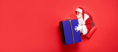 Santa Claus , the real one, comes out of the red studio background with a big gift. Christmas time.A lot of copy space.の写真素材