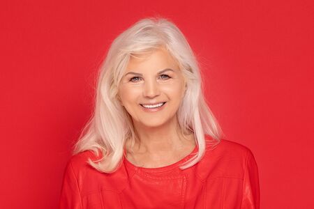 Beautiful senior woman with long grey hair posing in red fashionable clothes in studio, smiling to...