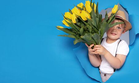 Young little caucasian kid holding yellow bouquet of flowers over blue background. Happy boy with smile. Mother's day.の写真素材
