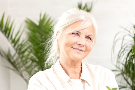 Portrait of smiling beautiful senior lady at home. Green houseplants on the background.の写真素材
