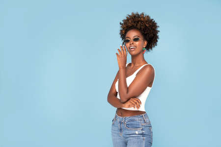 Fashionable young african girl with afro hairstyle posing in studio, wearing sunglasses and earrings. Blue pastel studio background.の写真素材