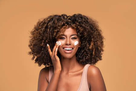 Young afro woman with moisturizer on face. Beautiful african girl applying a cream on cheek. Beauty girl with perfect and healthy skin.の写真素材