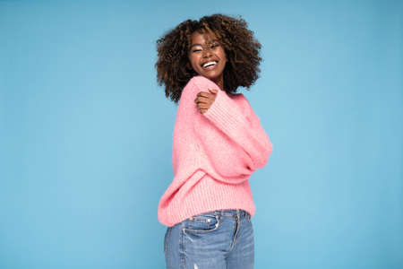 Happy smiling afro girl in pink sweater posing on blue studio background.