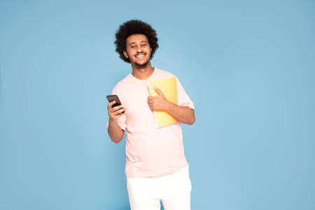 Cheerful young african guy in casual t-shirt holding mobile phone and notebook, smiling.