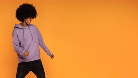 Happy man with afro hair wearing fashionable hoodie, dancing over orange background.