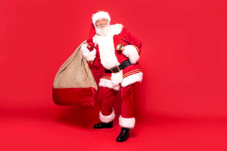 Happy, real, funny Santa Claus holding sack bag with Merry Christmas presents, delivering xmas gifts. Red background, a lot of copy space.の写真素材