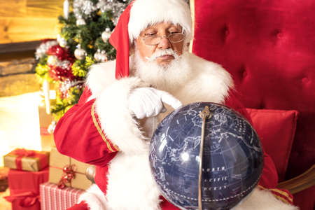 Real Santa Claus Sitting Near Christmas Tree and looking at the globe.