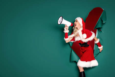 Beautiful blonde Santa girl in Christmas hat and costume shouting on megaphone.の写真素材