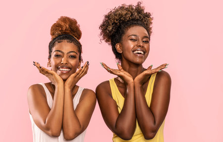 Happy overjoyed two afro women smile broadly, looking at camera, posing on pink pastel studio background.の写真素材