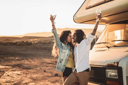 Happy beautiful hipster couple toasting with wine at sunset, standing near old camper. Travel, nomadic, real people lifestyle and emotions concept.の写真素材