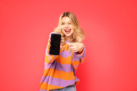 Happy beautiful young blonde woman posing over colorful studio background, pointing at mobile phone...