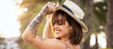Young beautiful happy brunette woman wearing summer hat, smiling charmingly. Girl with tattoos. Real people emotions. Summer, good vibes.の写真素材