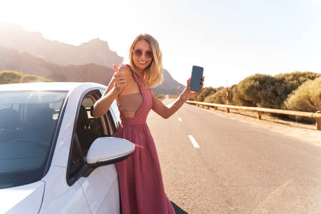 Happy blonde woman showing smartphone and showing thumb up, standing near the rental car. Summer vibes. Road trip.の写真素材