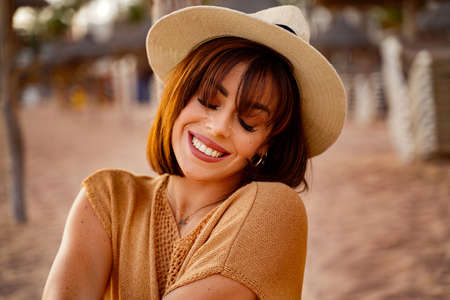 Closeup portrait of smiling young latino woman at the beach. Cheerful young girl enjoying a day on the sea shore at sunset, wearing summer hat.の写真素材