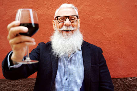 Handsome senior man with white beard and mustache toasting with red wine, looking at camera with big...