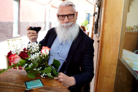 Elegant hipster senior man on a date in restaurant, holding red wine glass and flowers, smiling and looking to the camera. Leisure time. Outdoor bar.の写真素材