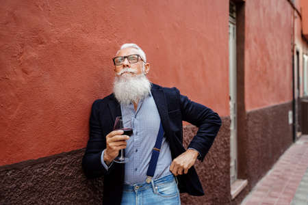 Handsome fashionable senior man with white beard and mustache holding glass of red wine and thinking about something. Outdoor photo. Elegant pensioner posing.の写真素材