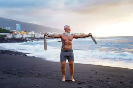 Age is just a number. In a healthy body, healthy mind. Senior man with white stylish beard exercising on the beach. Summer vacation workout. Sunset light. Elderly people healthy lifestyle on a retirement concept.の写真素材