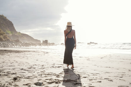 Beautiful romantic woman walking on the empty beach during sunset, wearing summer hat, looking at the ocean. Holidays island vibes.の写真素材