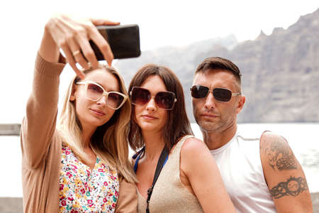 Three smiling friends taking selfie photos at digital camera of smartphone. Summer time. One man and...