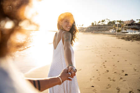 Lovely smiling beautiful woman holding hand of her boyfriend, looking at him, walking on the beach during sunset. Summer island vibes. Love is in the air. Real people lifestyle.の写真素材