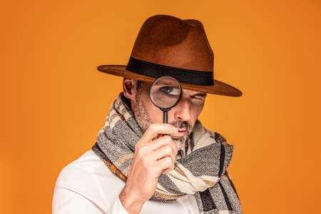 Male detective with magnifying glass, wearing fashionable scarf and brown hat, posing on yellow studio background. Portrait of handsome man with beard.の写真素材