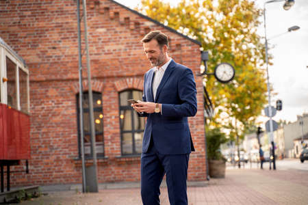 Photo of handsome smiling businessman using mobile phone outdoor on the city street. Thoughtful businessman in elegant suit talking by smartphone.の写真素材