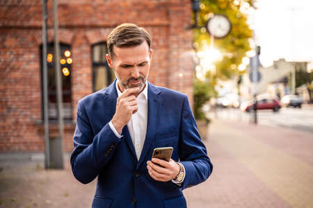 Photo of handsome businessman using mobile phone outdoor on the city street. Thoughtful businessman in elegant suit working on smartphone.の写真素材