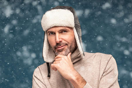 Portrait of handsome man wearing winter cap, posing over snow flakes, looking at camera. Closeup face of cheerful guy wearing warm clothes.の写真素材