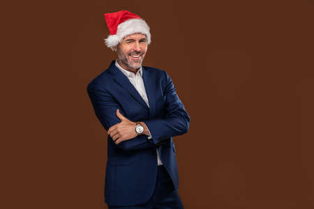 Happy handsome businessman wearing santa claus cap winks to the camera on brown studio background and smiling.の写真素材