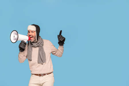 Smiling handsome man in winter clothes holding megaphone and screaming about a big sale, isolated on blue pastel studio background.の写真素材