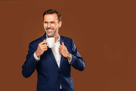 Successful handsome business man in classic fashionable suit posing on brown background in studio, drinking hot coffee. Achievement career wealth business concept.の写真素材