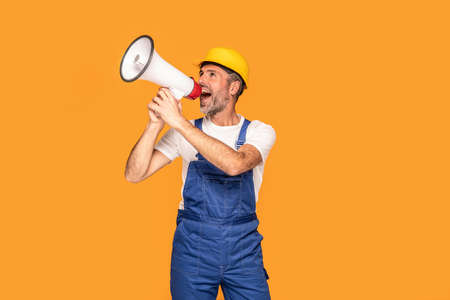 Handsome happy construction worker shouting loud by megaphone with smile and positive emotions on his face. Marketing or sales concept. Yellow studio background.の写真素材