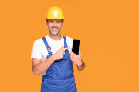 Smiling Construction Worker Showing Cellphone Empty Screen to the camera. Yellow studio background.の写真素材
