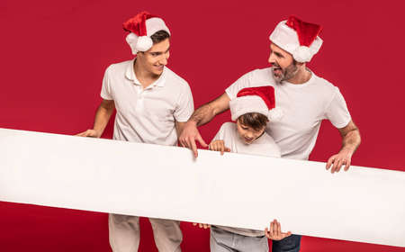 Male part of the family in a festive atmosphere. A dad and two sons are holding a white empty board, posing on a red studio background, wearing Santa Claus hats, smiling and celebrating christmas time.の写真素材