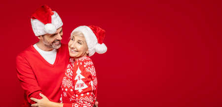 A loving, happy mature couple in santa hats posing against a red background in the studio, hugging and smiling heartily. Christmas time. Wife and husband in xmas sweaters. A lot of copy space.の写真素材