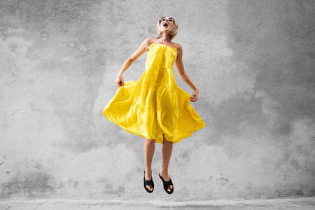 Happy laughing blonde woman jumping in yellow dress over gray wall. A lot of copy space. Traveler. Real people emotions.の写真素材