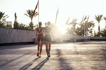 Tired beautiful fit blonde women taking a break, happy after outdoor workout. Two friends walking together. Back view. Sport. Hobby. Girls with ideal slim bodies.の写真素材