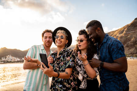 Group of multiracial happy young friends laughing, taking selfie with mobile phone, having fun together on the beach. Multi ethnic friendship concept. Summer time. Traveler. Real people emotions.の写真素材