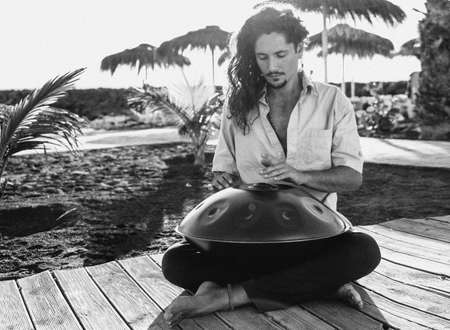 Handsome italian man practicing native music on a hang drum, a traditional drumming instrument played with hands. Leisure time on the beach. Sunset time.の写真素材
