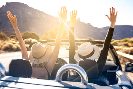 Dreams come true! Two happy young girls driving cabrio car during vacation road trip in mountains, making memories and having fun together. Back view. Hands up. Freedom concept. Happiness. Tourist. Wanderlust. Real people emotions.の写真素材