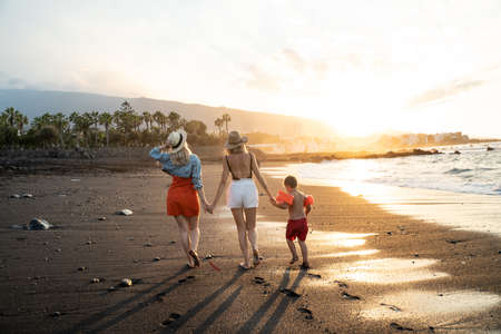 Happy homosexual family with kid walking on the beach during sunset, holding hands. Back view. Tropical vacation. Lesbians with son.の写真素材