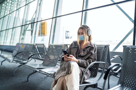 Caucasian woman in mask using smartphone and headphones at airport terminal sitting and waiting for an airplane, watching something on social media. Traveler. Tourist. flight transport.の写真素材