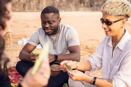 Group of happy multiracial friends having fun together on the beach, playing cards on picnic,...
