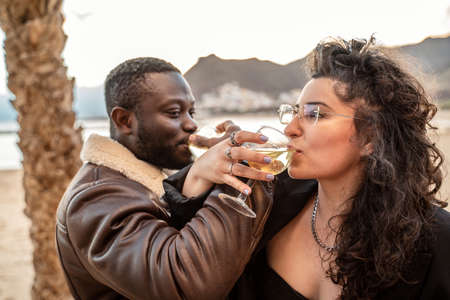 Beautiful multiracial couple enjoying glass of champagne on the beach at sunset. Love. Honeymoon. Emotions. People celebrating proposal.の写真素材