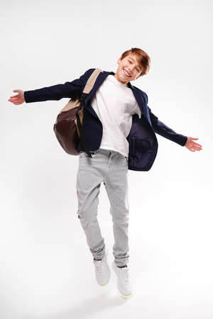 Young energetic handsome happy school boy in fashionable clothes jumping with backpack over studio background, smiling to the camera. Real people emotions. Full photo.の写真素材