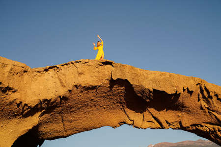 Young woman in yellow maxi dress in emotional dance outdoor in the nature on the sandstone. Blue...