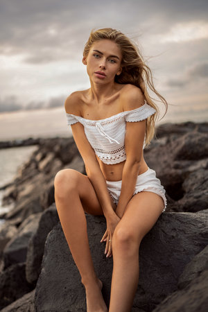 Caucasian blonde young woman relaxing on the rocky beach. Sensual girl looking at the camera, sitting on the rocks. Summer vibes.の写真素材