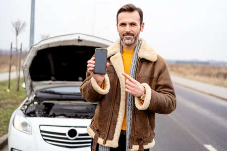 A man by the side of the road is standing in front of a broken car, pointing on an empty mobile phone screen, smiling to the camera. Application with insurance. Engine problem. A lot of copy space.の写真素材